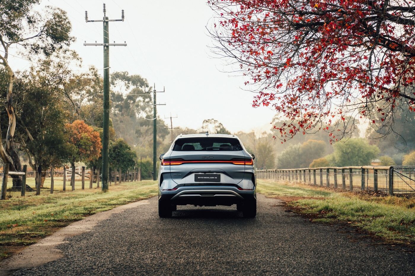An electric SUV driving away down a quiet, tree-lined country road in autumn